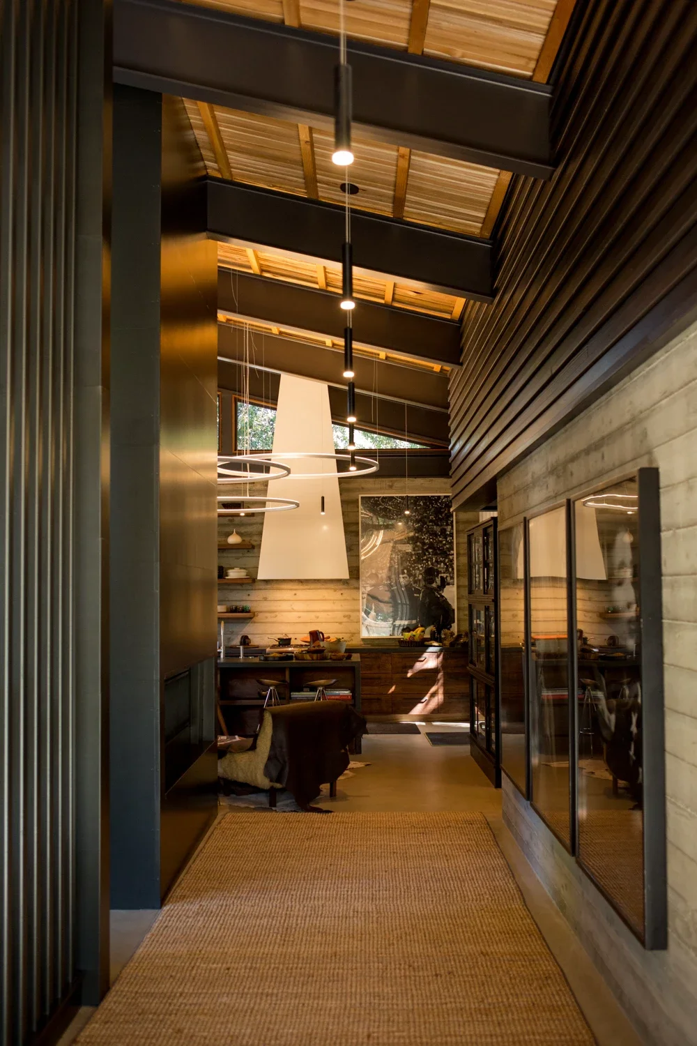 Interior view of a modern kitchen with wood-paneled and black walls, sloped wooden ceiling, hanging pendant lights, and a large window reflecting trees and a person taking the photo.
