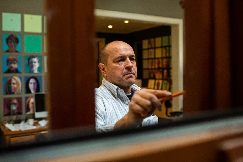 Ray Turner wearing a striped shirt, is drawing on an easel in a room with framed pictures of people's faces on the wall behind him.
