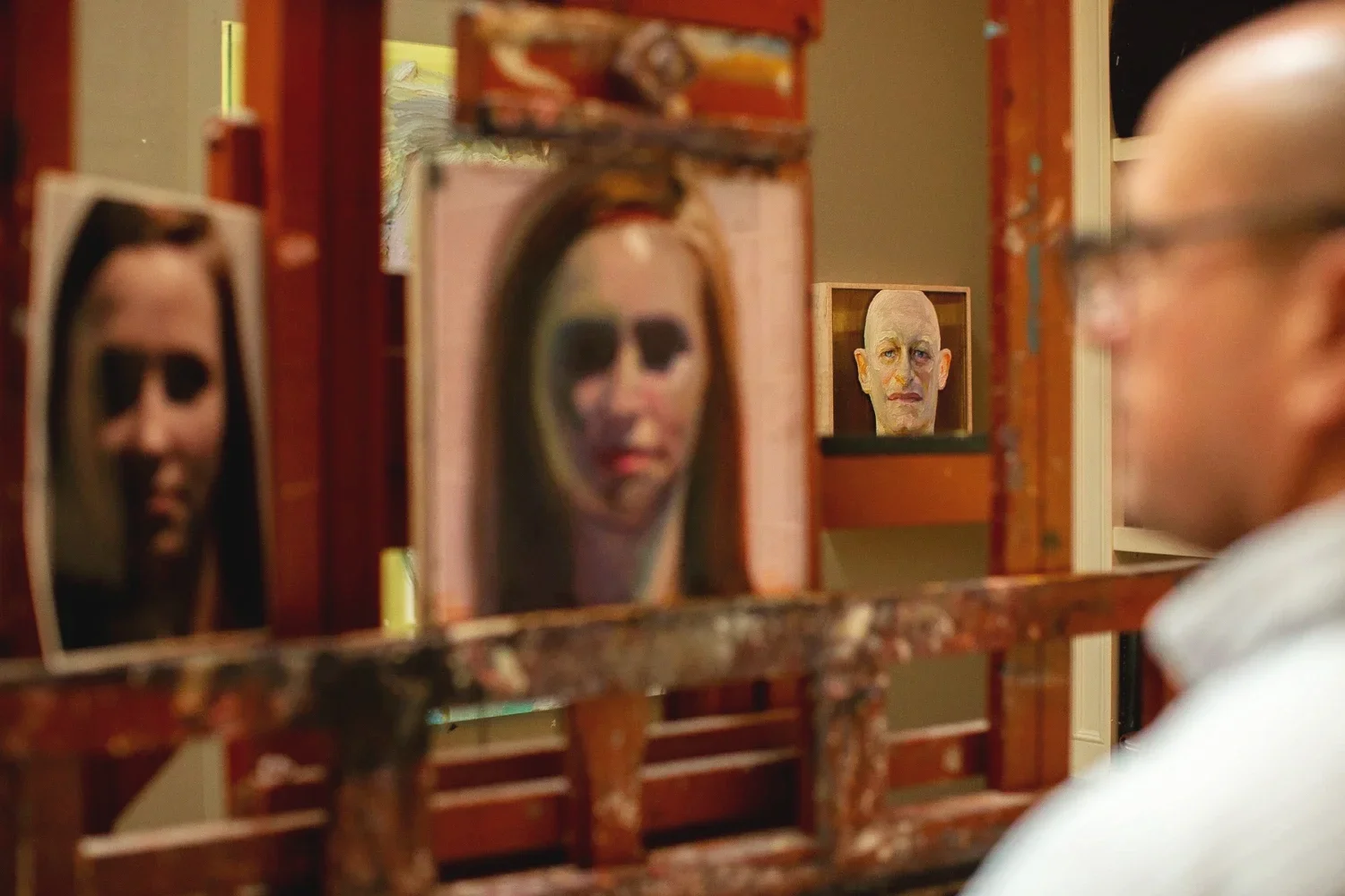 Person looking at his paintings of human faces, with multiple framed face images in the background.