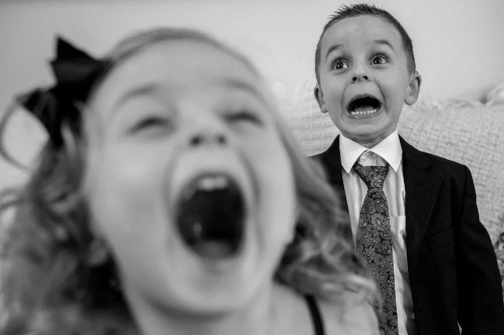 Two children, a girl in the foreground with a bow in her hair and a boy in a suit and tie, both screaming with wide-open mouths and expressive faces.