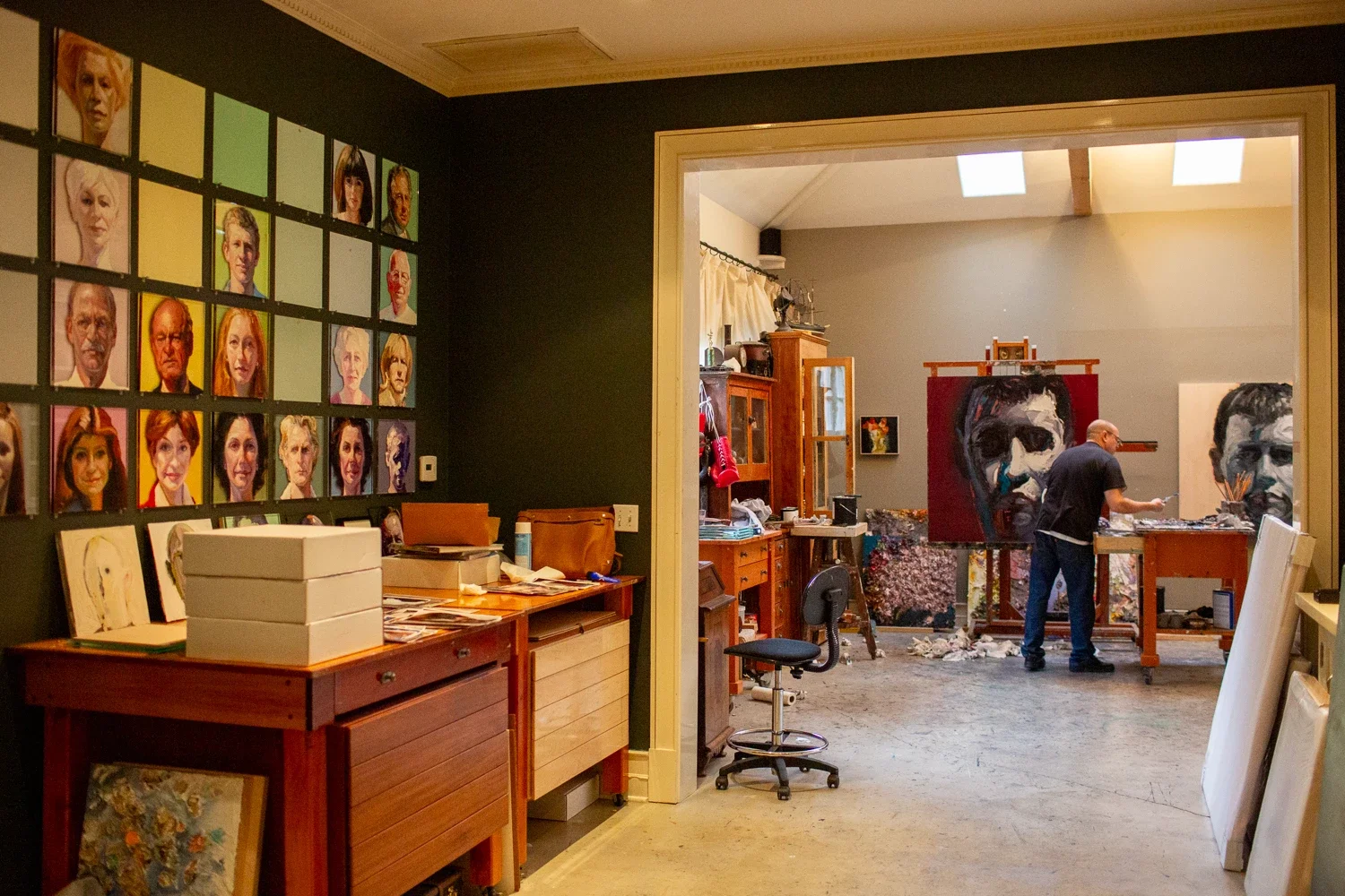 Artist's studio with portraits on the wall, a workspace with an artist painting a large portrait, and various art supplies and furniture.