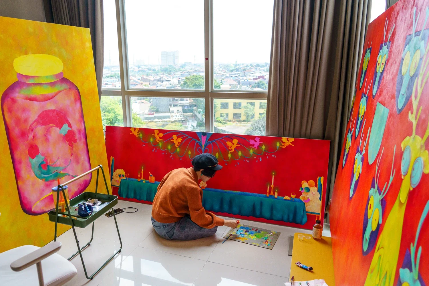 Person sitting on the floor painting colorful abstract artwork in a room with a large window. Three large, vibrant paintings are visible around the person, and a small table with art supplies is nearby.