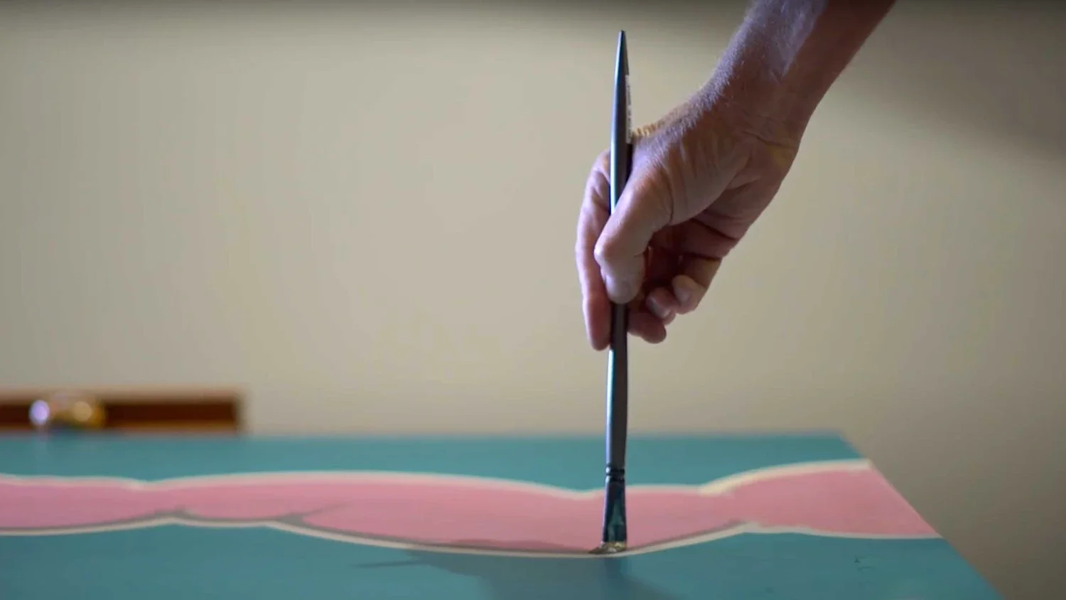 A hand holding a paintbrush, painting a pink and white abstract design on a canvas.