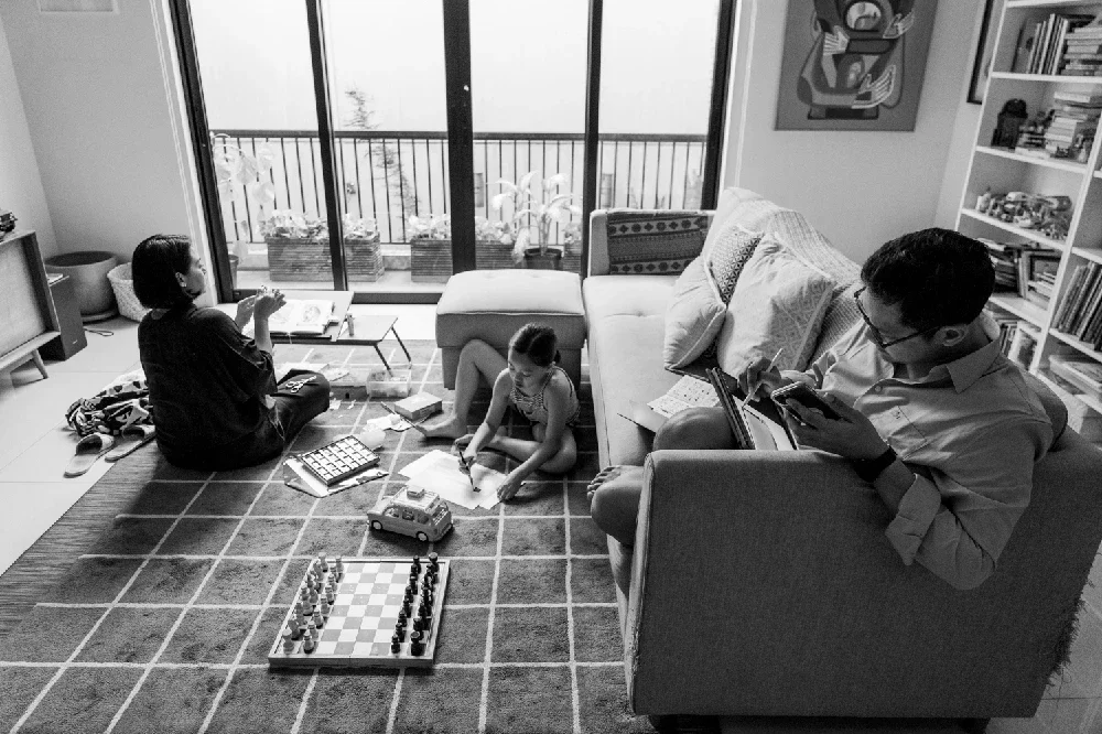 A family in a living room, with a woman sitting on a mat with a child playing with a toy truck, a man on a couch reading a book, and a girl drawing on paper on the floor, near large sliding glass doors leading to a balcony.