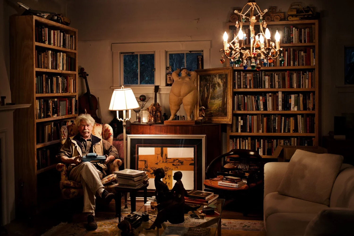 Master builder Frank Gallagher sitting in a floral armchair, holding a vintage tin toy car in a cozy living room filled with bookshelves, a chandelier, and various artworks.