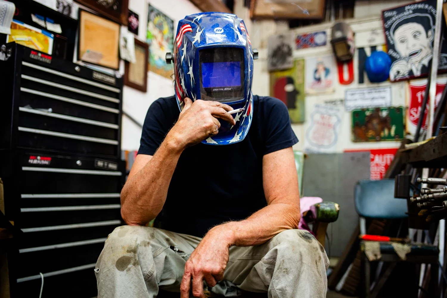 A man with a welding mask holding a cigarette sitting down in the artistic metal shop
