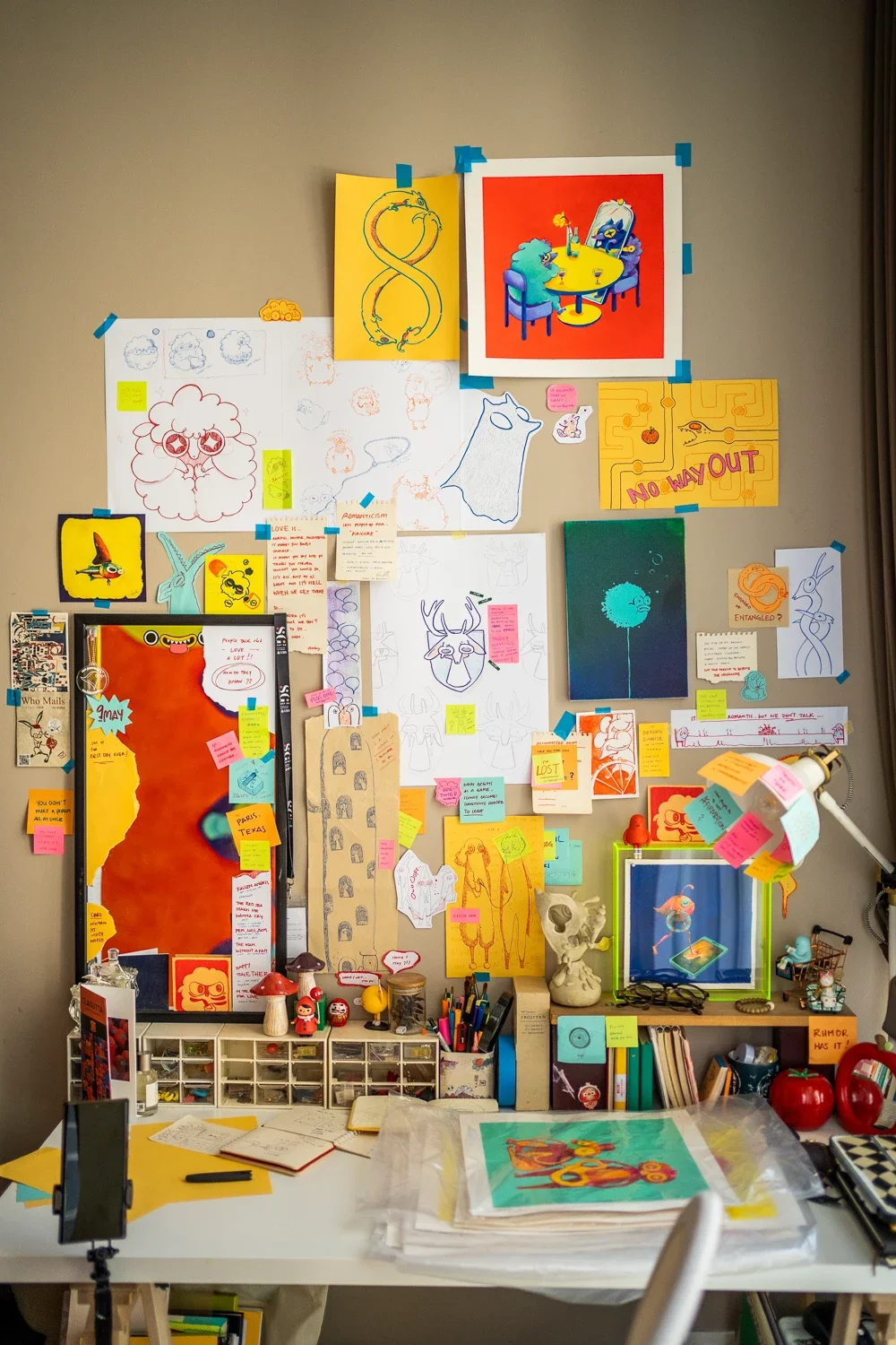A wall decorated with colorful illustrations, sketches, sticky notes, and artwork, with a desk below holding various art supplies, books, and decorative items.