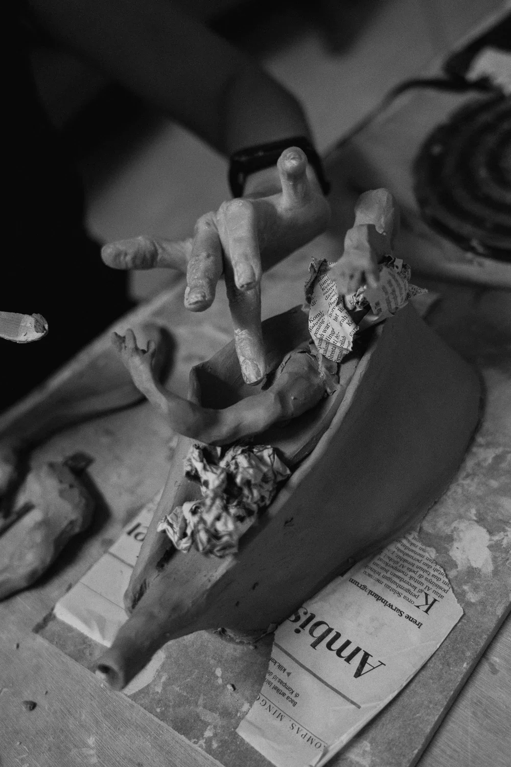 Close-up of a sculpture in progress, featuring a human hand with fingers splayed, on a table with crumpled newspaper and tools visible, in black and white.
