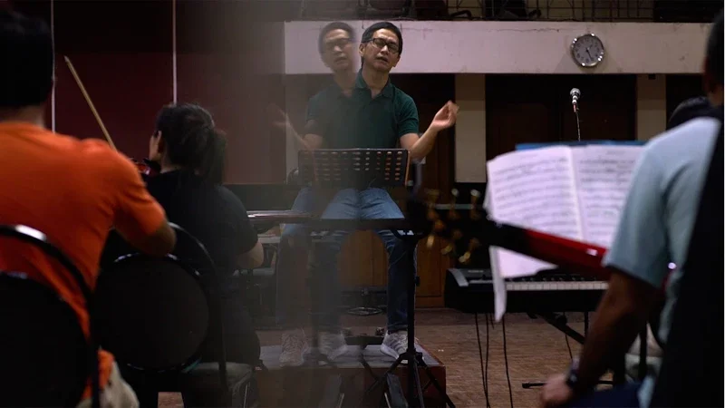 Music conductor leading an orchestra in a rehearsal, with musicians playing instruments and sheet music in front.
