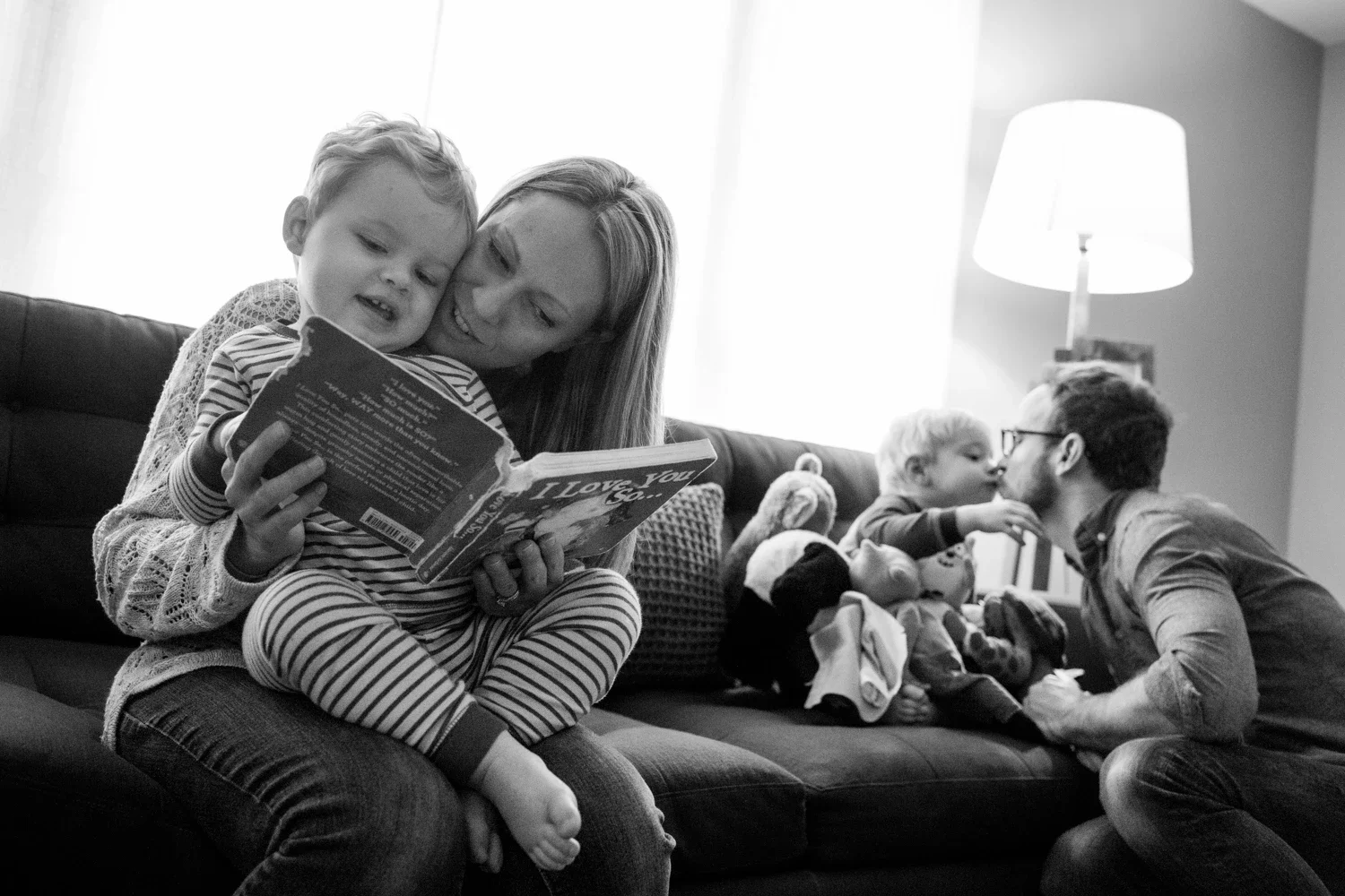 A family sitting on a couch reading and playing with their children in a cozy living room, black and white photograph.