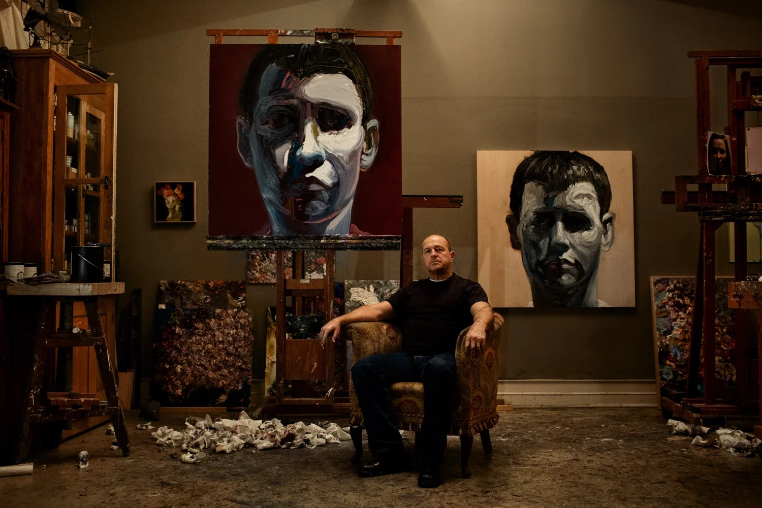 Ray Turner seated in his art studio surrounded by large portrait paintings of a face, with paint-covered canvases and crumpled paper on the floor.