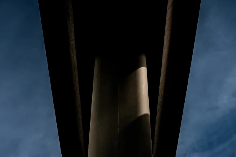 Looking up at the underside of a bridge or overpass, showing supporting pillars and beams against a blue sky with clouds.