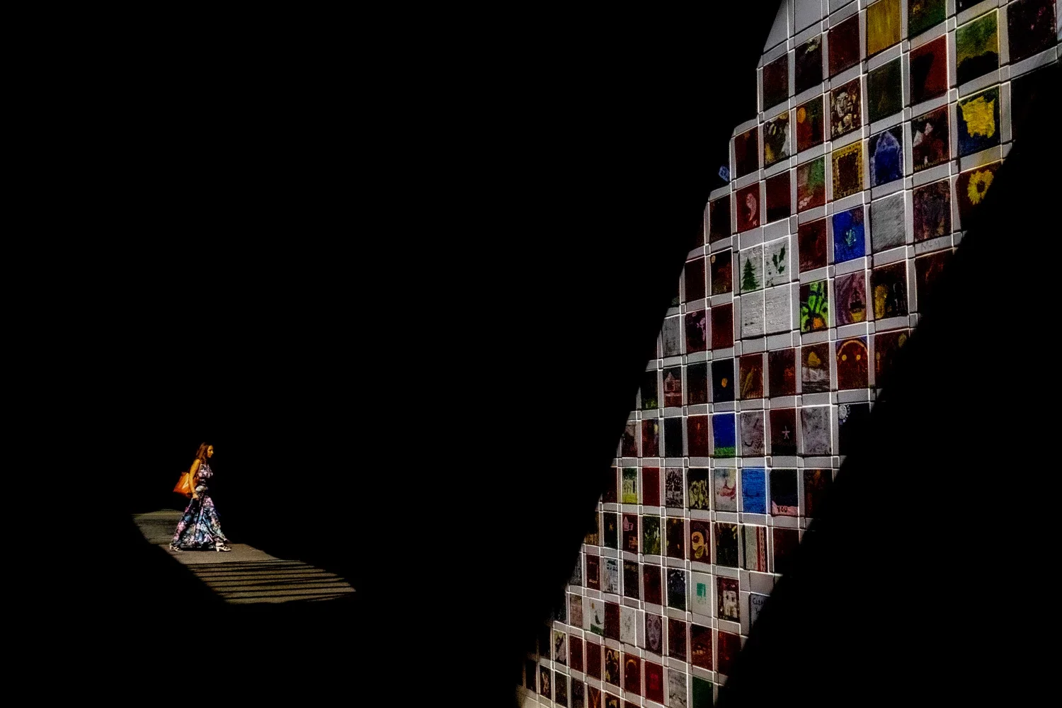 A woman walking on the street wearing a long, colorful dress with a bag over her shoulder, seen through a dark opening with a large mural of loaded grocery bags on a glass building in the background.