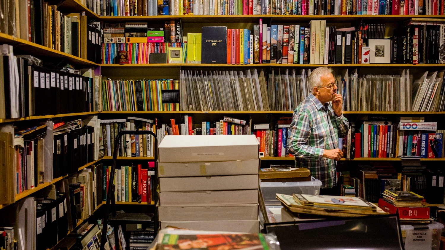 An older man with glasses wearing a plaid shirt standing in a bookstore or library, talking on a phone, surrounded by shelves filled with books and records.