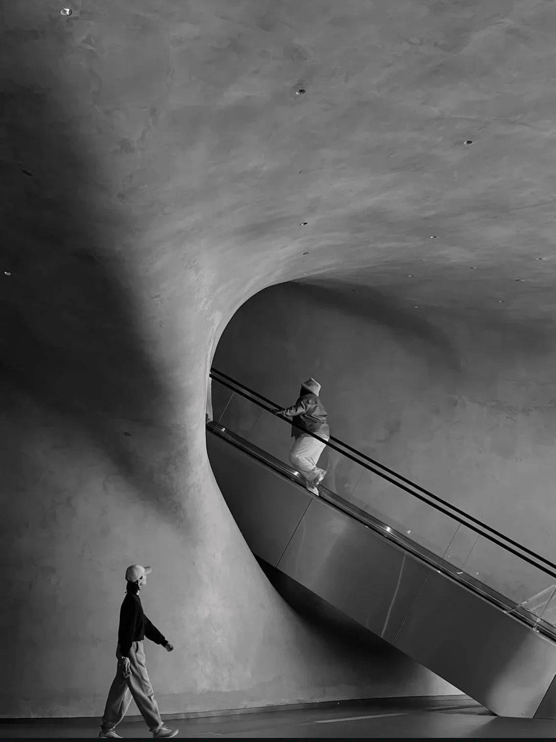 People in a modern, curved architectural space with an escalator and concrete walls.