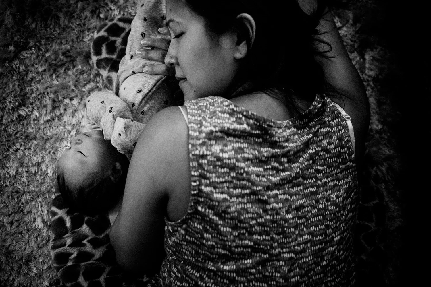 A woman and a young child are lying on a textured surface, with the woman resting her head near the child's head. The woman has closed eyes and appears peaceful, while the child is sleeping soundly. The scene is in black and white.