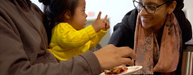 A young girl in a yellow sweater making hand gestures and smiling while talking to two women, one in a gray jacket and the other in glasses and a scarf, who are smiling and sitting at a table with food.
