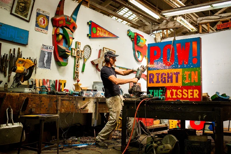 David Buckingham in a workshop filled with colorful signs and art pieces, holding a power grinder, working on a large sign that reads 'POW! RIGHT IN THE KISSER' in bold, multicolored letters.