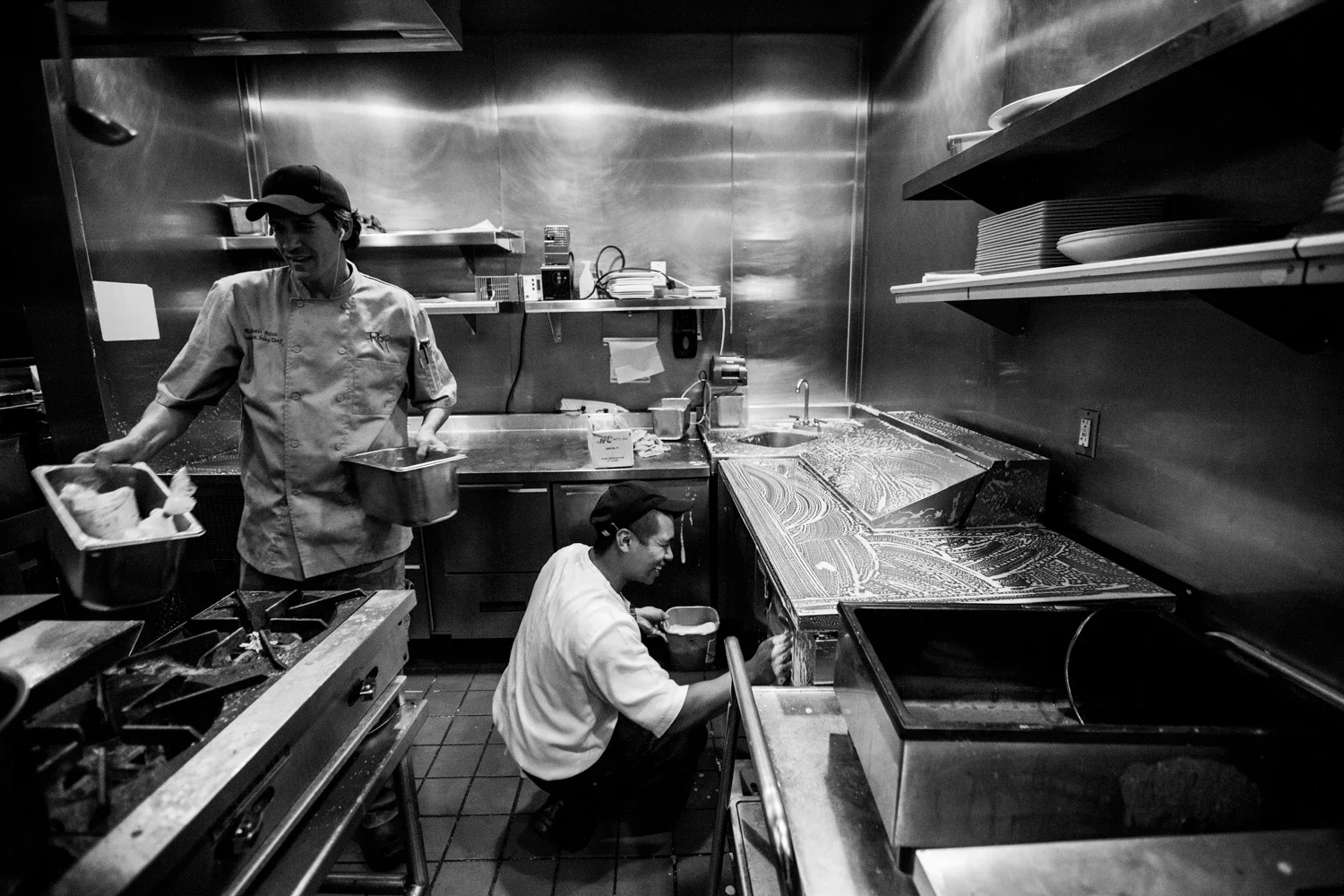 Two chefs smiling and making jokes as they perform thorough cleanup in the kitchen at the end of the night