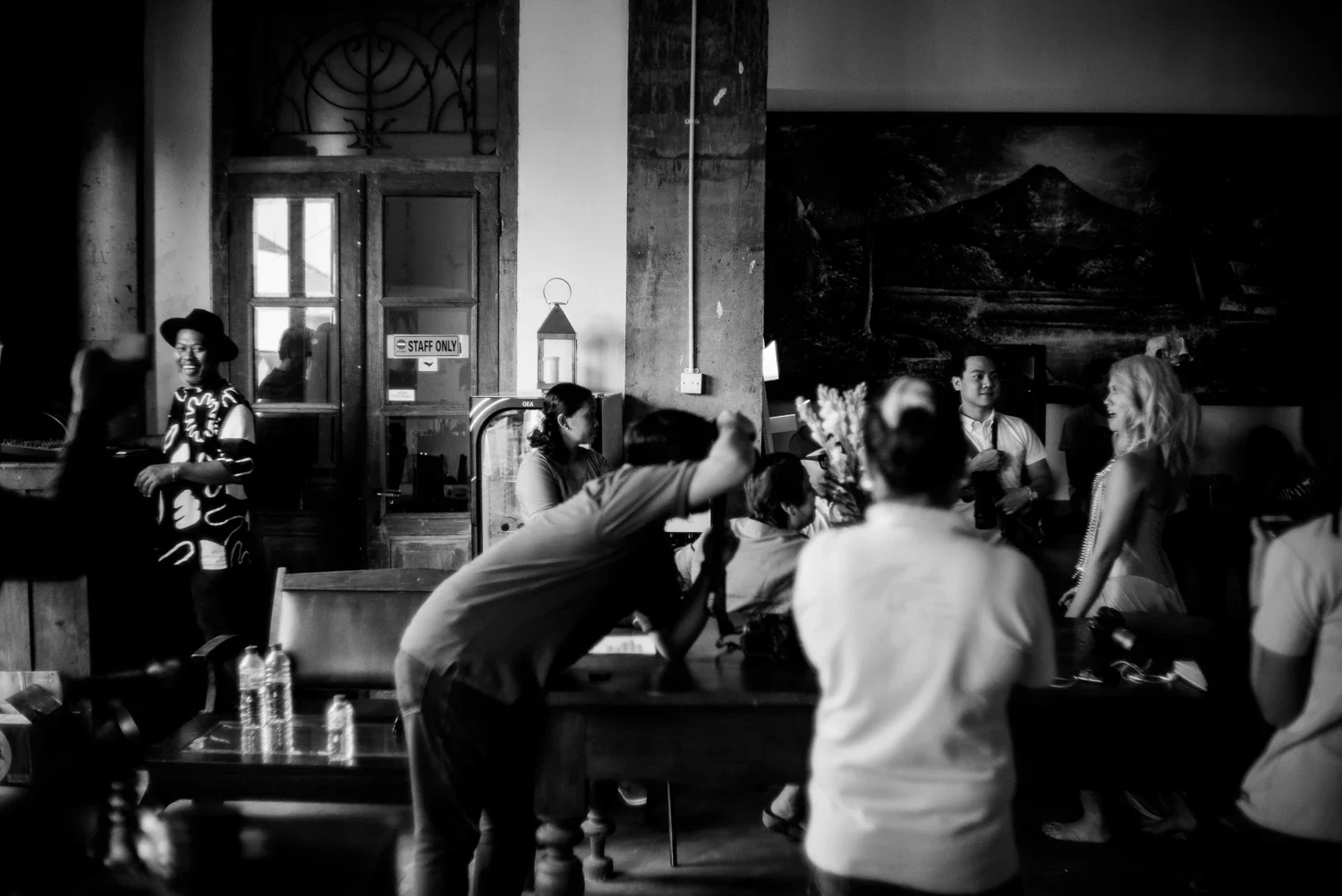 A black and white photo of people socializing indoors at a restaurant or café. There are several women and men, some seated at tables and others standing, engaging in conversation. A woman with blond hair is talking to a man, and a woman in a patterned dress and hat is smiling near the door. The background features a large mountain painting on the wall, wooden doors, and a small sign that says 'STAFF ONLY.'
