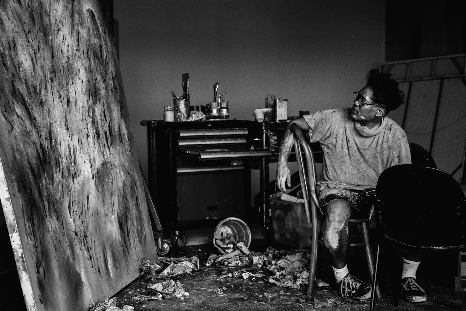 Anton Ismael sitting in a cluttered art studio, looking at a large abstract painting on the wall, with various art supplies and tools on a nearby table.