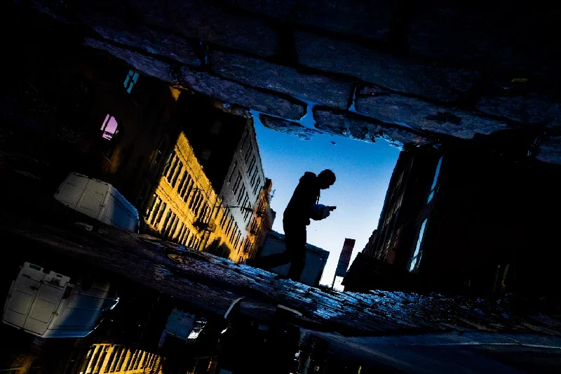 Silhouette of a person walking on a city street at dusk, viewed from below through a large puddle reflecting the sky and surrounding buildings.