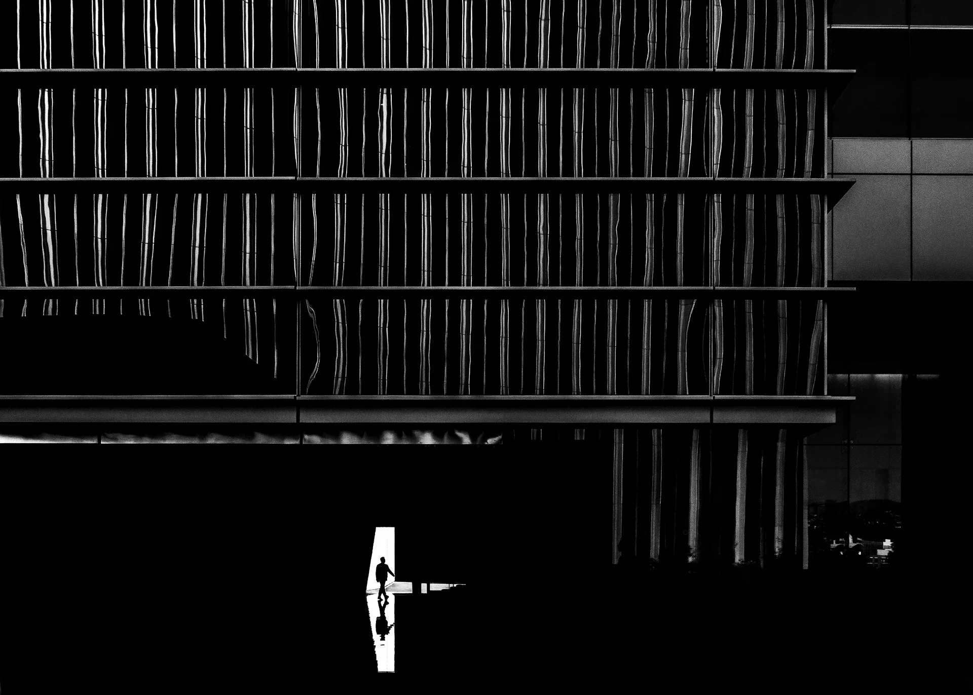 A person walking through a dark passageway beneath a modern building with patterned glass windows.