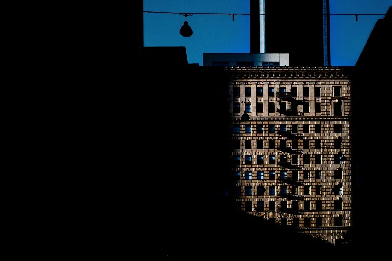 A tall building with multiple windows, partly in shadow, against a deep blue sky with a silhouette of a hanging streetlight and a rooftop structure.