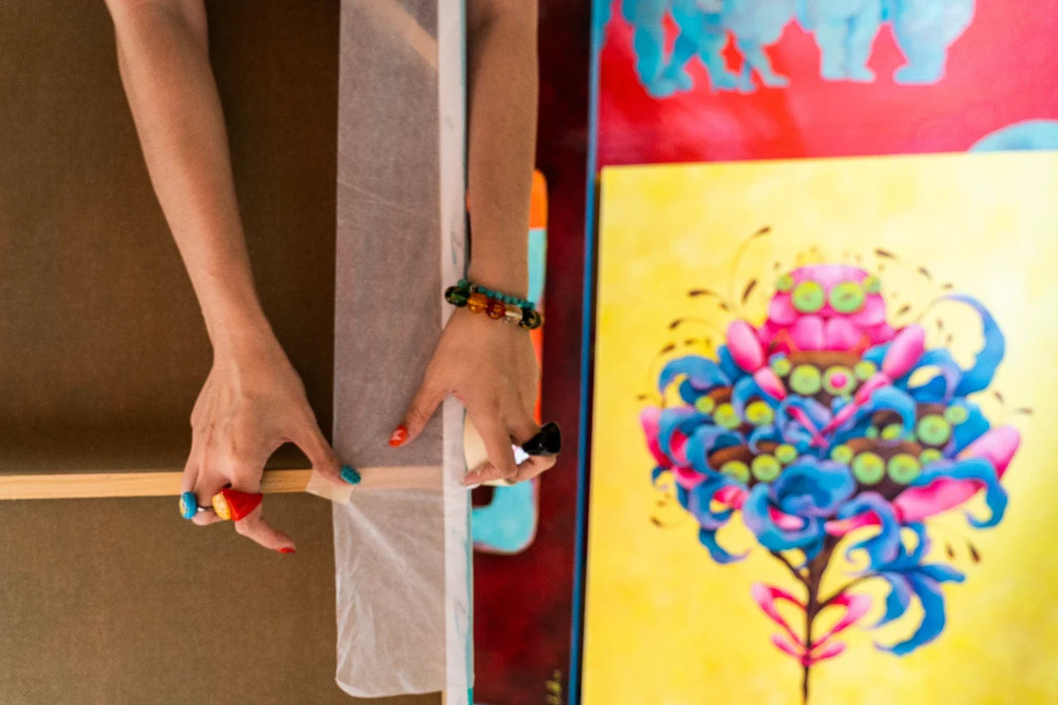 Person's hands with colorful rings and bracelets, cutting paper on a table with vibrant artwork in the background.
