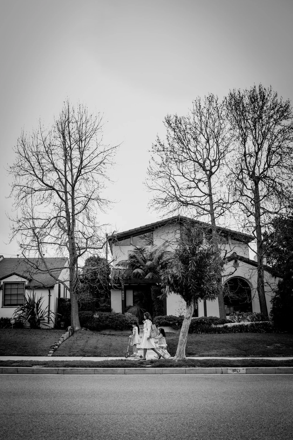 A black and white photo of children walking on the sidewalk near a house with tall, leafless trees in front, in a suburban neighborhood.