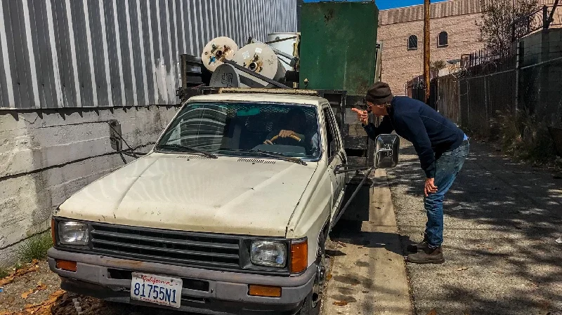 Metal Sculptor David Buckingham leaning over to talk to a driver of a truck containing old metal cabinets in a downtown Los Angeles alley.