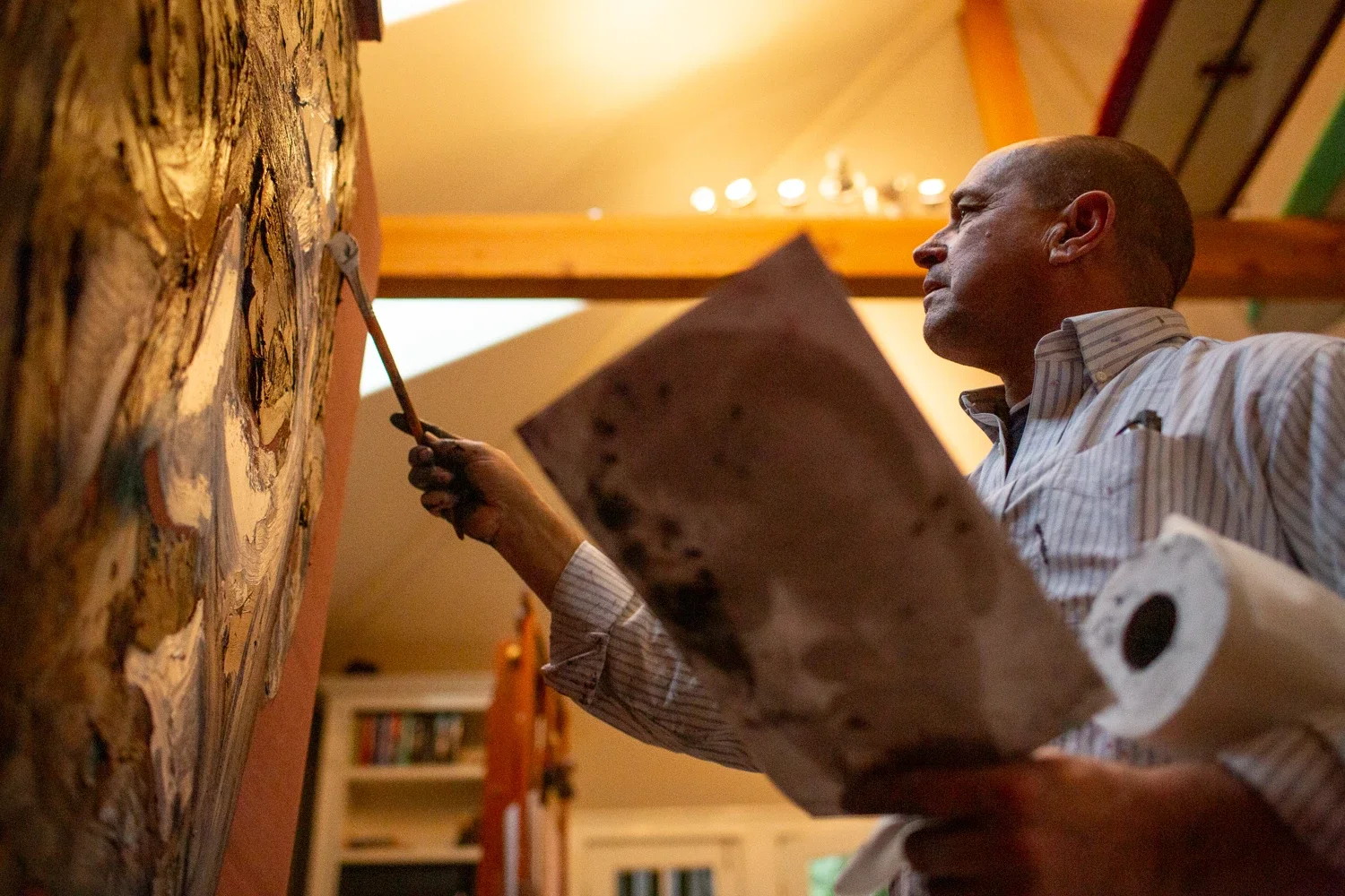 A man in a striped shirt painting a detailed picture on a large canvas inside a room.