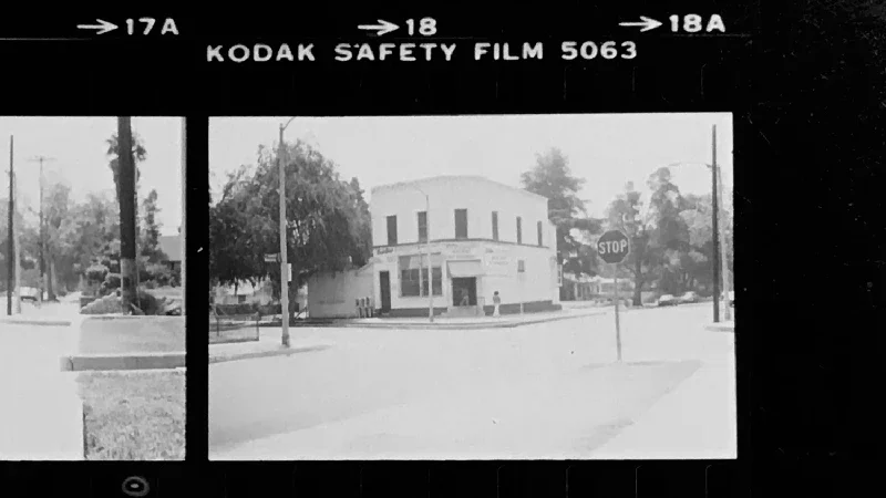 Black and white photo of a downtown street corner with a stop sign, a two-story building, trees, and power lines. The photo is taken through Kodak safety film frame.