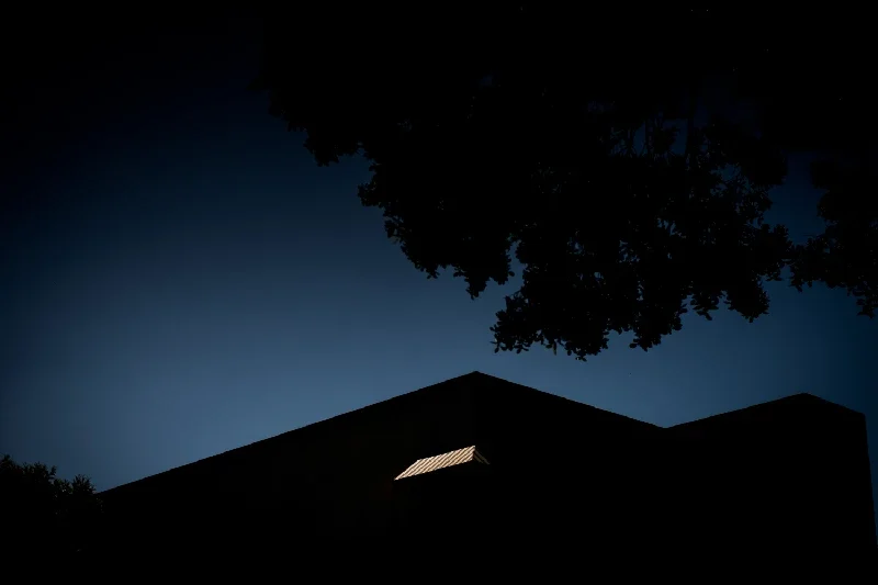 Silhouette of a house with a small window on the roof, surrounded by trees, against a dark blue evening sky.
