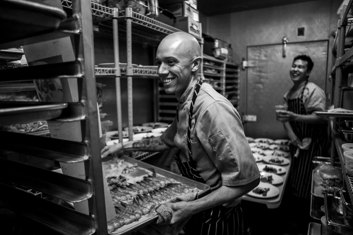 Two chefs working in a crowded room prepping food for a big group while smiling and making jokes