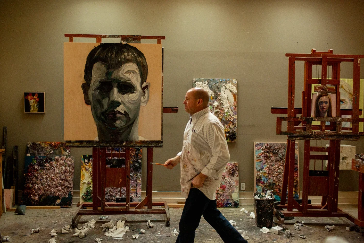 An artist in a studio, walking past partially finished portrait paintings of people on easels and the wall, with paint splatters on the floor and canvases.