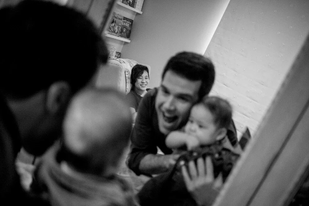 A man playfully holds a young girl in a corner, while two adults and a baby observe and smile in a cozy indoor setting.