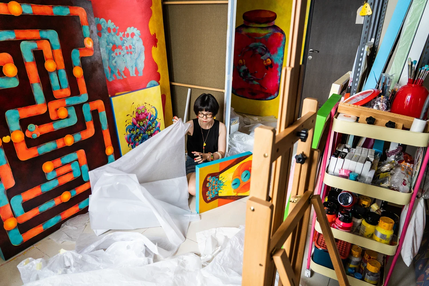An artist with short black hair and glasses arranging paintings in a brightly colored art studio. Various paintings and art supplies surround her, with a cluttered workspace including shelves with paint bottles and brushes.