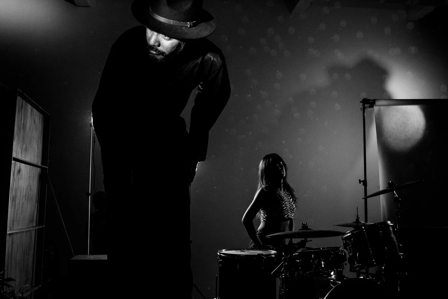 Black and white photo of a man with a hat and beard looking down, and a woman with long hair playing drums in a dimly lit studio or stage with a backdrop and a light source creating shadows.