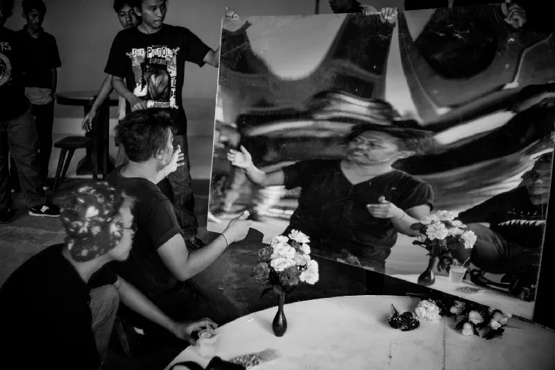 Group of people gathered around visual artist Anton Ismael in front of a large mirror. A woman is in front of the mirror, adjusting her appearance. Flowers in vases are on the table.