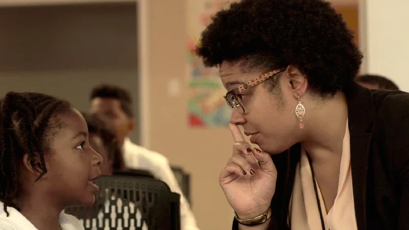 A woman with glasses and earrings leans forward, listening attentively to a young girl with braided hair, in a classroom setting.