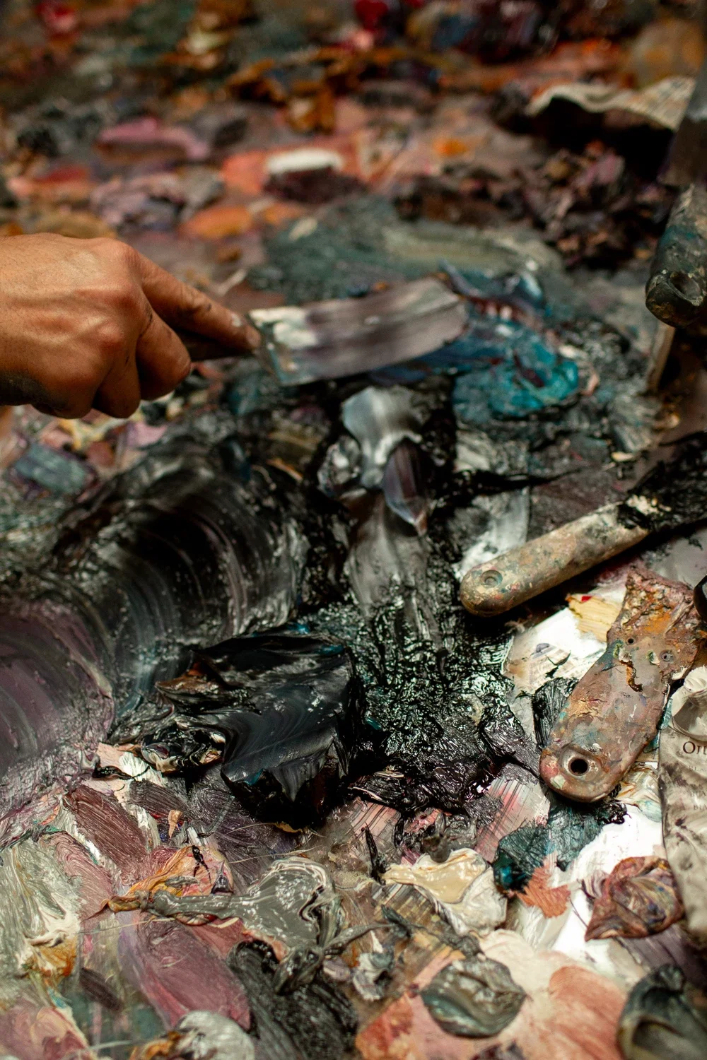 A hand holding a palette knife scrapes thick oil paint from an artist's palette, with mixed paint colors and a paintbrush in the scene.