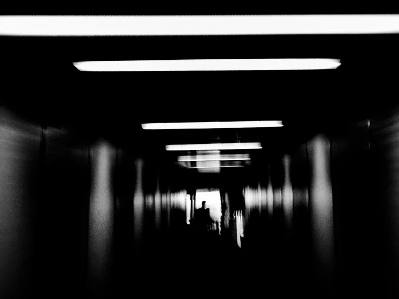 Silhouette of a person sitting in a chair at the end of a dark corridor with bright overhead lights.