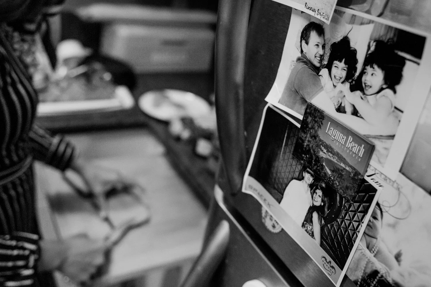 Black and white photograph showing photographs and postcards attached to a wooden surface, with a blurred background of a person's hand holding chopsticks and eating food.