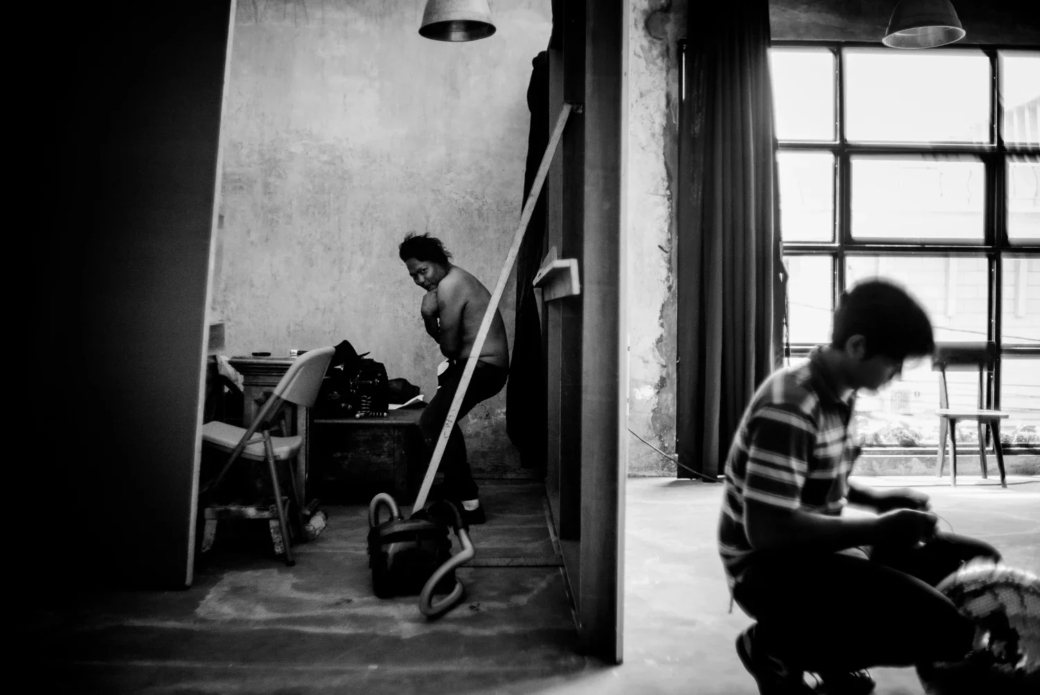 Black and white photo of two people in a room with large windows. One woman is standing, partially undressed, at a desk, and a young man is sitting on the floor, looking at something.