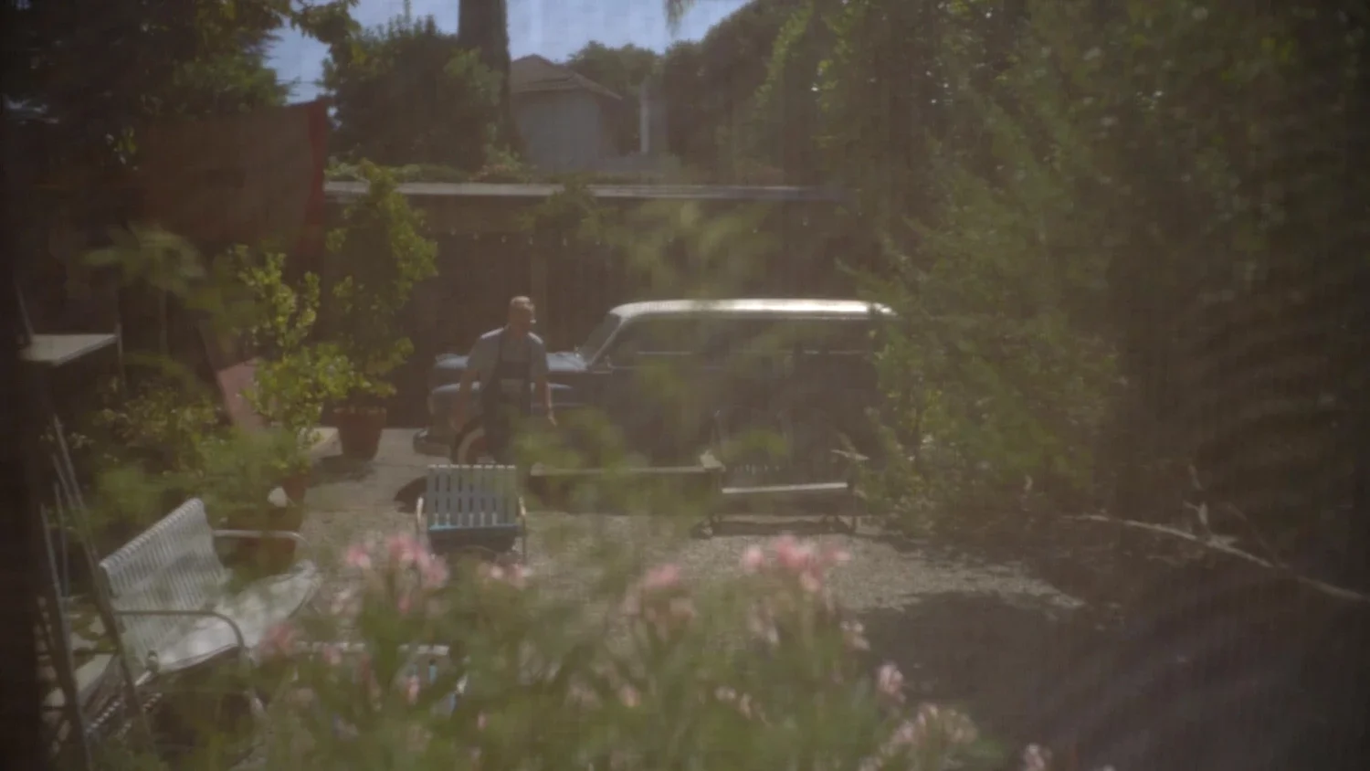 A person walking in a backyard with a vintage car, garden benches, and trees, viewed through a window with reflections.