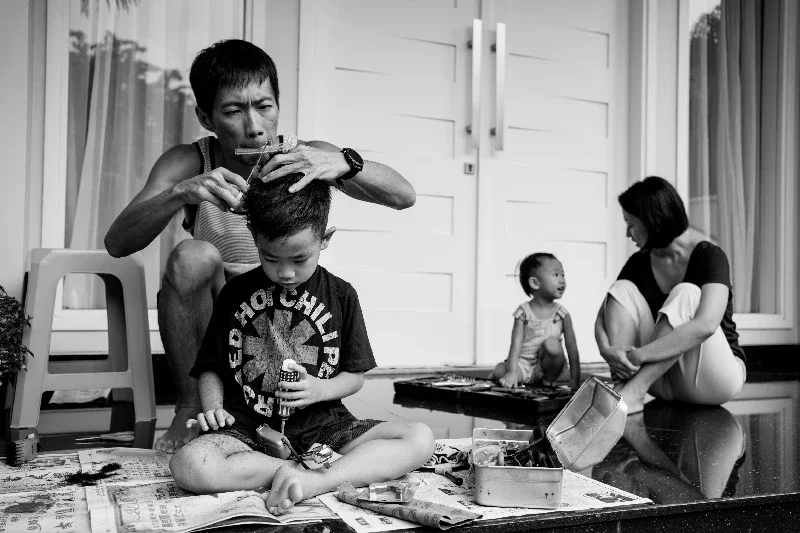 A father cutting the hair of his boy at the front porch of his house while his wife and daughter are talking in the background