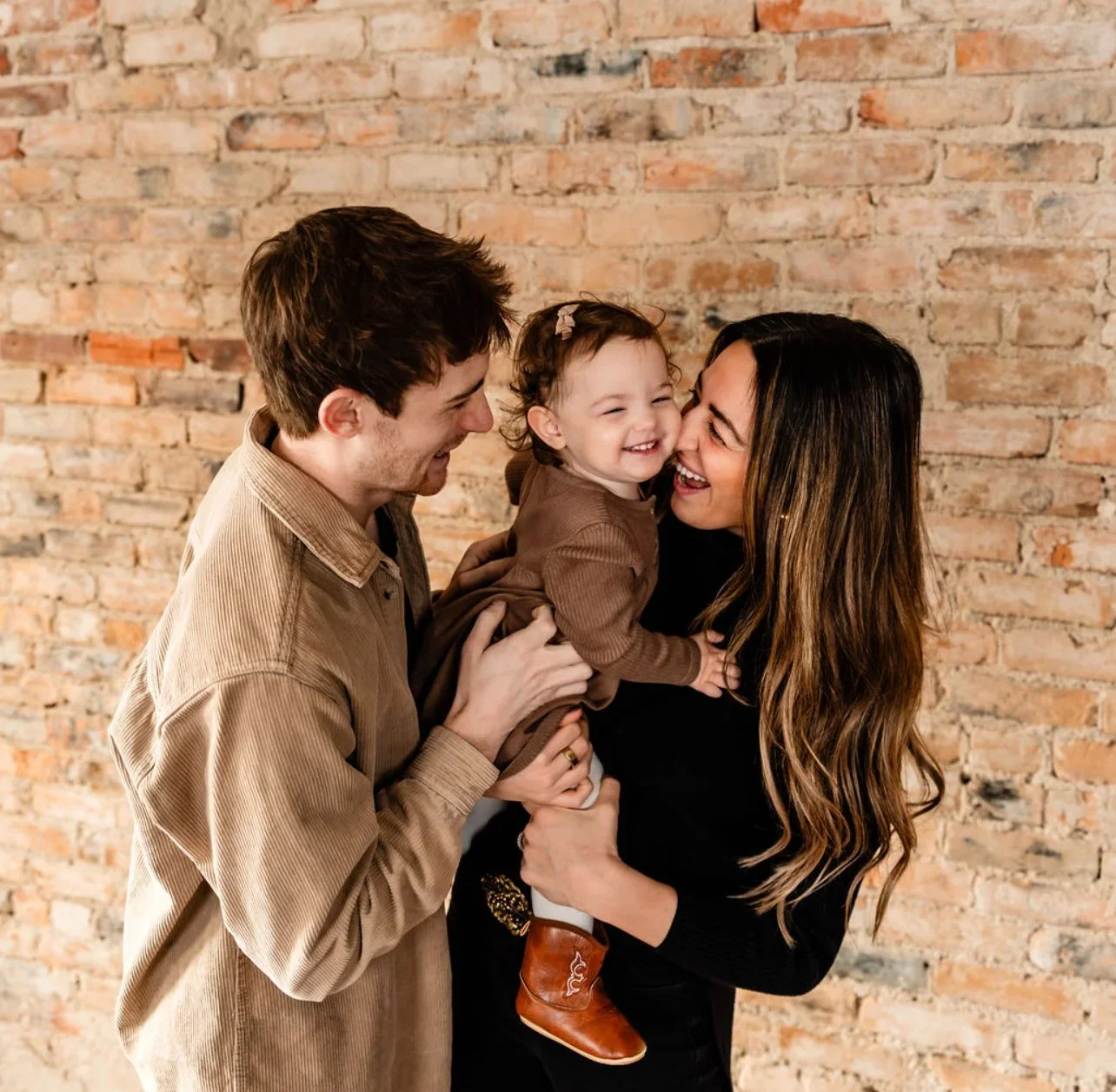 A man, woman, and baby girl laughing together in front of a brick wall.