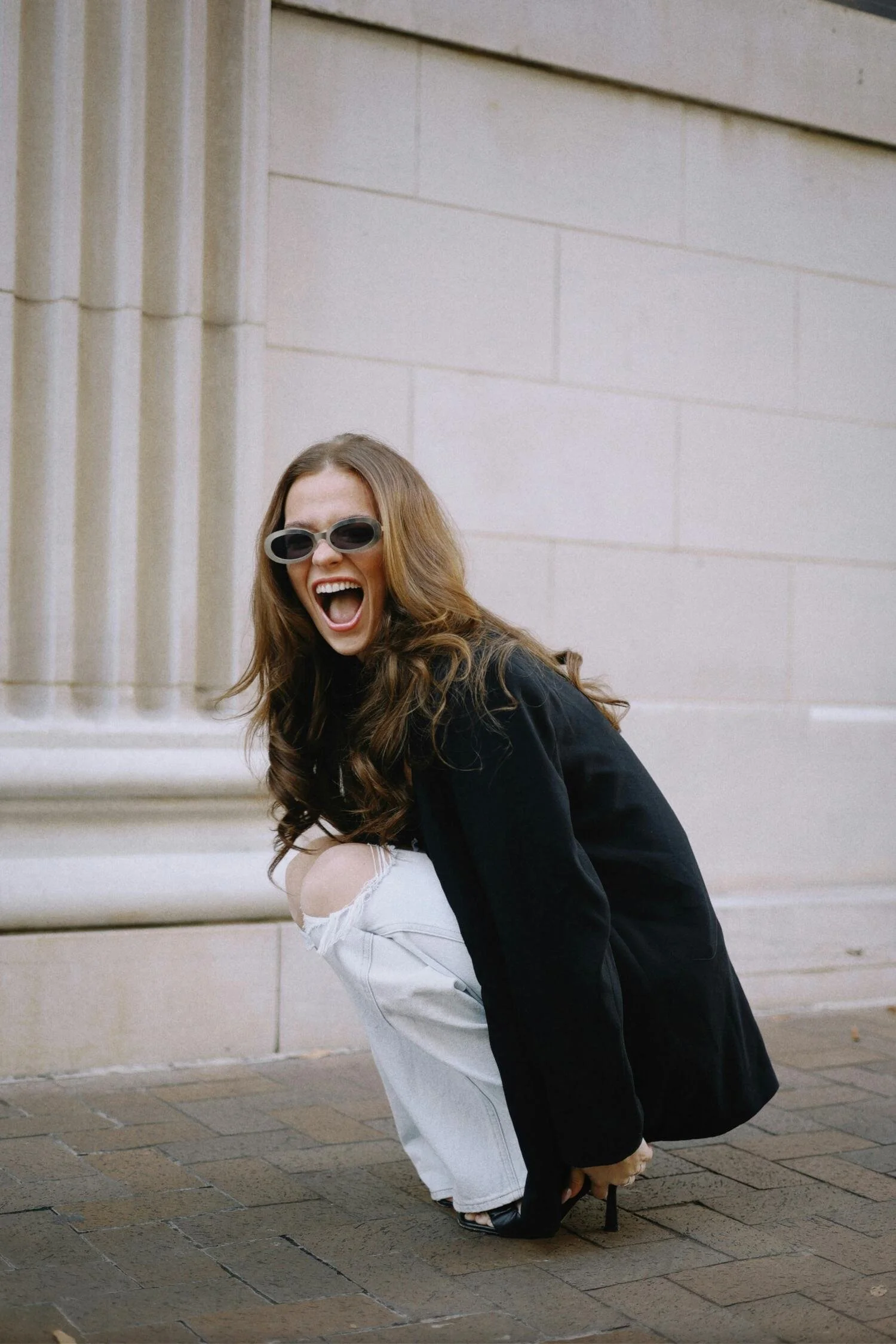A young woman with long brown hair, wearing black sunglasses, black jacket, white ripped jeans, and high heels, is crouching on the sidewalk in front of a beige stone building, smiling and appearing joyful.