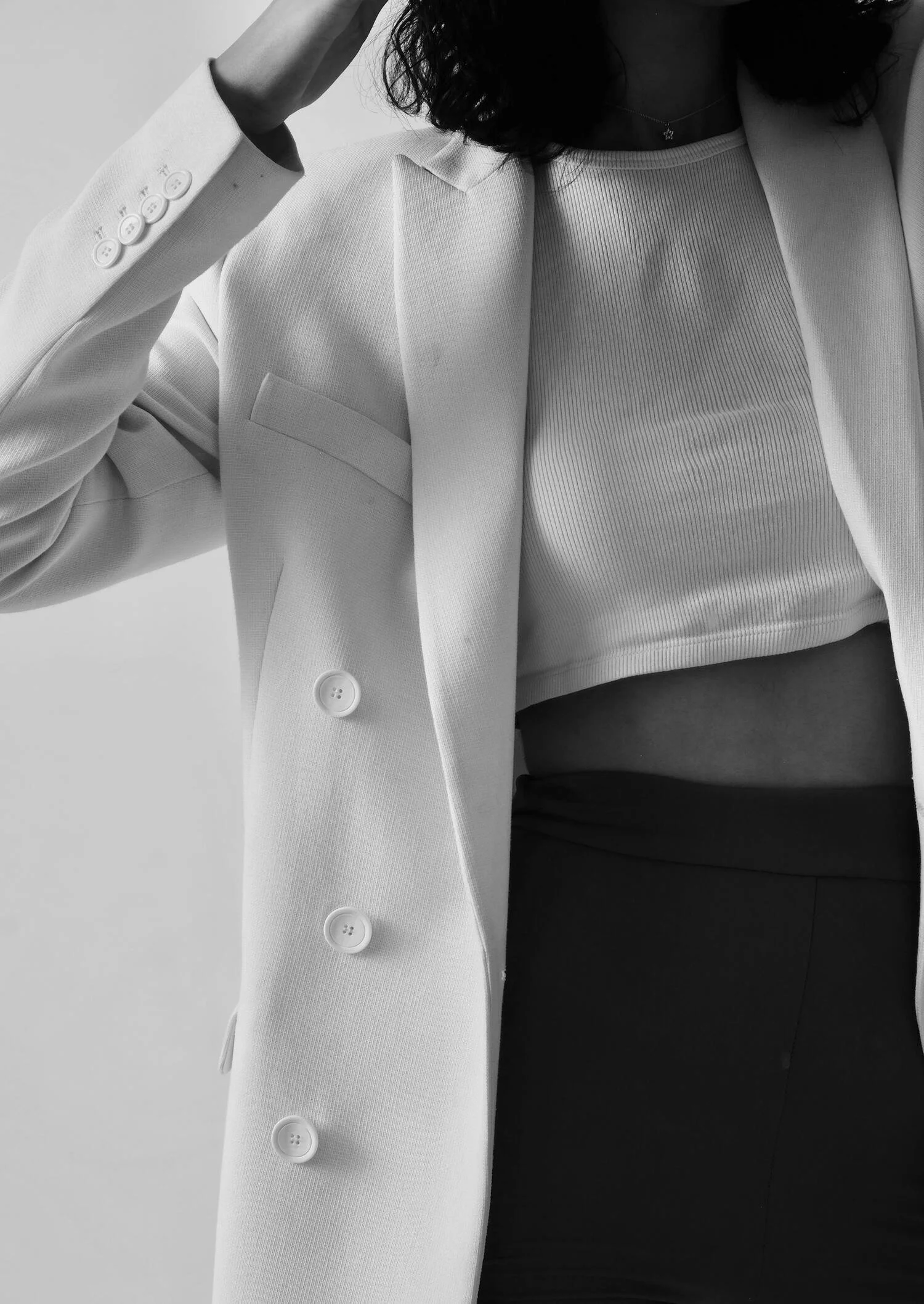 A person dressed in a white blazer, ribbed top, and dark pants, with their hand touching their head, black and white photography.